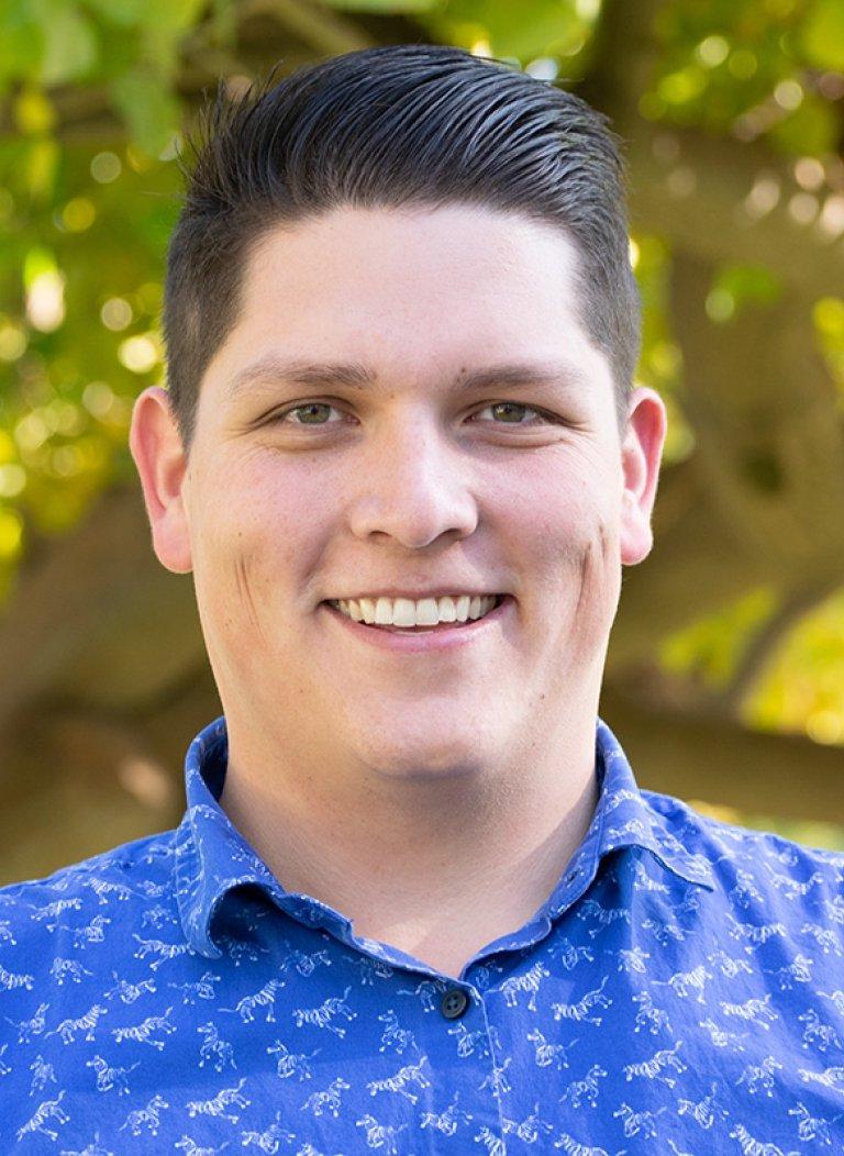 Man with dark hair, smiling, wearing a blue-collared shirt, standing in front of greenery.